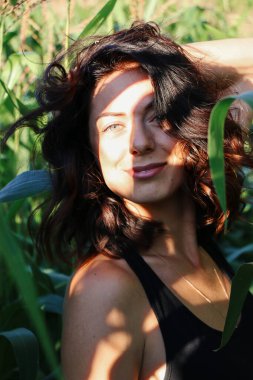 Yazın mısır yaprakları içinde güzel esmer bir kız, doğadaki bir kadının sanat portresi. Kameraya bakarak kahkahalar atmak, seçici keskinlik, açık iş gölgesi doğal ışık.