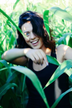 Yazın mısır yaprakları içinde güzel esmer bir kız, doğadaki bir kadının sanat portresi. Kameraya bakarak kahkahalar atmak, seçici keskinlik, açık iş gölgesi doğal ışık.