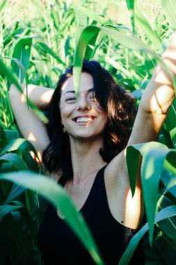 Yazın mısır yaprakları içinde güzel esmer bir kız, doğadaki bir kadının sanat portresi. Kameraya bakarak kahkahalar atmak, seçici keskinlik, açık iş gölgesi doğal ışık.