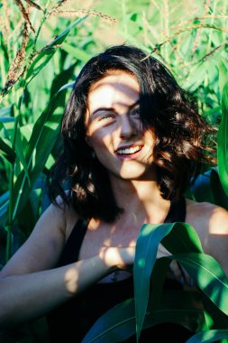 Yazın mısır yaprakları içinde güzel esmer bir kız, doğadaki bir kadının sanat portresi. Kameraya bakarak kahkahalar atmak, seçici keskinlik, açık iş gölgesi doğal ışık.