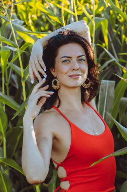 Kırmızı elbiseli güzel esmer kız yaz mısır yaprakları, doğadaki bir kadının sanat portresi. Kameraya bakarak kahkahalar atmak, seçici keskinlik, açık iş gölgesi doğal ışık.