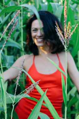 Kırmızı elbiseli güzel esmer kız yaz mısır yaprakları, doğadaki bir kadının sanat portresi. Kameraya bakarak kahkahalar atmak, seçici keskinlik, açık iş gölgesi doğal ışık.