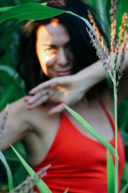 Kırmızı elbiseli güzel esmer kız yaz mısır yaprakları, doğadaki bir kadının sanat portresi. Kameraya bakarak kahkahalar atmak, seçici keskinlik, açık iş gölgesi doğal ışık.