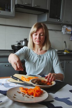 Yetişkin bir kadın, sarışın, ev mutfağında sakince yemek hazırlar, tuvayı kaydırır, yiyeceğe dokunur, yana ya da kameraya bakar. Doğal ışık altında basit bir ev işi..