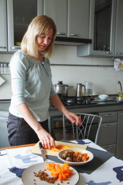 Yetişkin bir kadın, sarışın, ev mutfağında sakince yemek hazırlar, tuvayı kaydırır, yiyeceğe dokunur, yana ya da kameraya bakar. Doğal ışık altında basit bir ev işi..