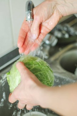 Yetişkin bir kadın, sarışın, sakince mutfakta yemek hazırlıyor, elinde yeşil lahana salıncağını tutuyor, yana ya da kameraya bakıyor. Doğal ışık altında basit bir ev işi..