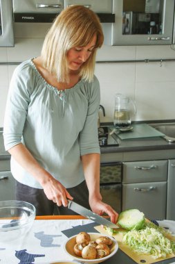 Yetişkin, güzel bir kadın, sarışın, ev mutfağında sakince yemek hazırlıyor, mutfakta yeşil lahana kesiyor. Doğal ışık altında basit bir ev işi..
