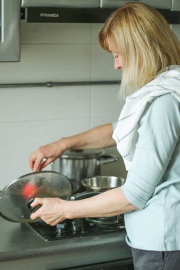Evin mutfağında elinde cam kapakla gaz sobasının yanında duran ve mantar kızartan sarışın bir kadının dikiz görüntüsü. Doğal ışık basit bir evcil süreçtir.