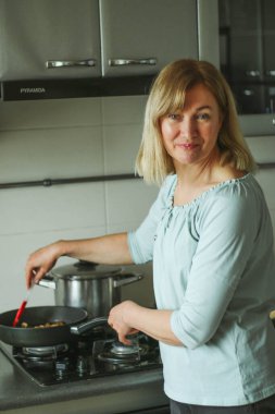 Evin mutfağında elinde cam kapakla gaz sobasının yanında duran ve mantar kızartan sarışın bir kadının dikiz görüntüsü. Doğal ışık basit bir evcil süreçtir.