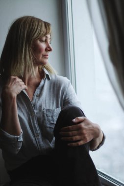 Pencerenin kenarında oturan yetişkin bir kadının portresi, dinleniyor, huzur ve yalnızlığın tadını çıkarıyor, pencereden dışarı bakıyor, doğal ışık,.