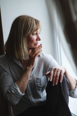 Pencerenin kenarında oturan yetişkin bir kadının portresi, dinleniyor, huzur ve yalnızlığın tadını çıkarıyor, pencereden dışarı bakıyor, doğal ışık,.