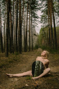 Sarışın, çıplak, genç, güzel bir kadın ormanda çimlerin üzerinde ayna tutuyor. Ekoloji konsepti, gezegene saygı, insan ve doğa arasındaki bağlantı..