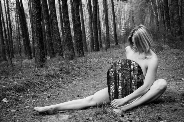 Blond nude young beautiful woman holding a mirror sitting on the grass in the forest. Ecology concept, respect for the planet, connection between man and nature.