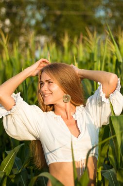 Yazın, batan güneşin ışığında, mısır tarlasında yeşil yaprakların arasında uzun kızıl saçlı güzel bir kadın..