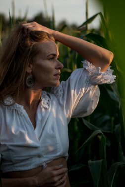 Yazın, batan güneşin ışığında, mısır tarlasında yeşil yaprakların arasında uzun kızıl saçlı güzel bir kadın..