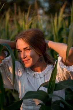 Yazın, batan güneşin ışığında, mısır tarlasında yeşil yaprakların arasında uzun kızıl saçlı güzel bir kadın..