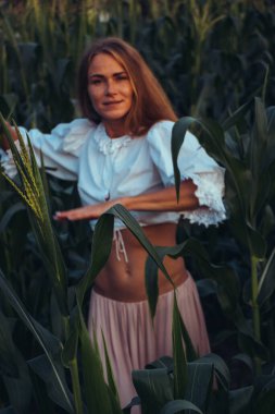 Yazın, batan güneşin ışığında, mısır tarlasında yeşil yaprakların arasında uzun kızıl saçlı güzel bir kadın..