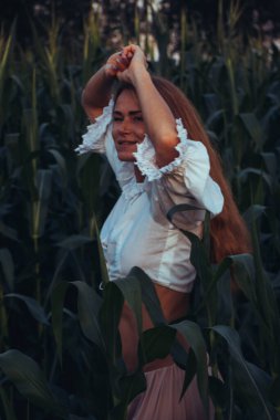 Yazın, batan güneşin ışığında, mısır tarlasında yeşil yaprakların arasında uzun kızıl saçlı güzel bir kadın..