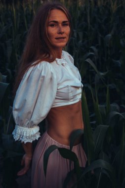 Yazın, batan güneşin ışığında, mısır tarlasında yeşil yaprakların arasında uzun kızıl saçlı güzel bir kadın..