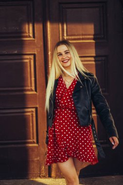 Kırmızı elbiseli ve siyah deri ceketli güzel bir kadının sokak portresi. Eski kahverengi bir kapının yanında duruyorum, doğal akşam gün batımı ışığı.