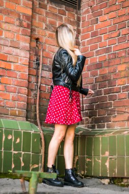 Kırmızı elbiseli ve siyah deri ceketli güzel bir kadının sokak portresi. Eski bir tuğla duvarın yanında duruyorum, doğal akşam ışığı.