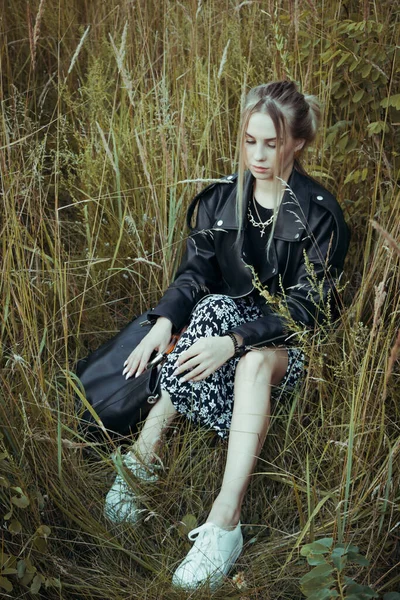 牧草地の背景に青い目の若い美しいブロンドの女性の肖像画 草の上の黒い服の中に座る自然の柔らかい夜の光 — ストック写真