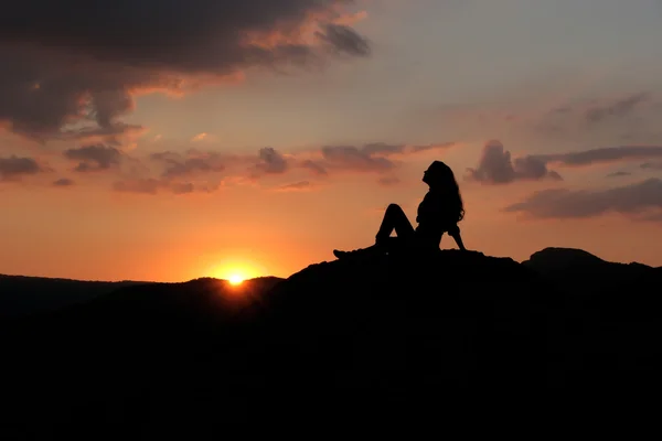 一个女孩坐在小山上落日的映衬下的剪影 — 图库照片