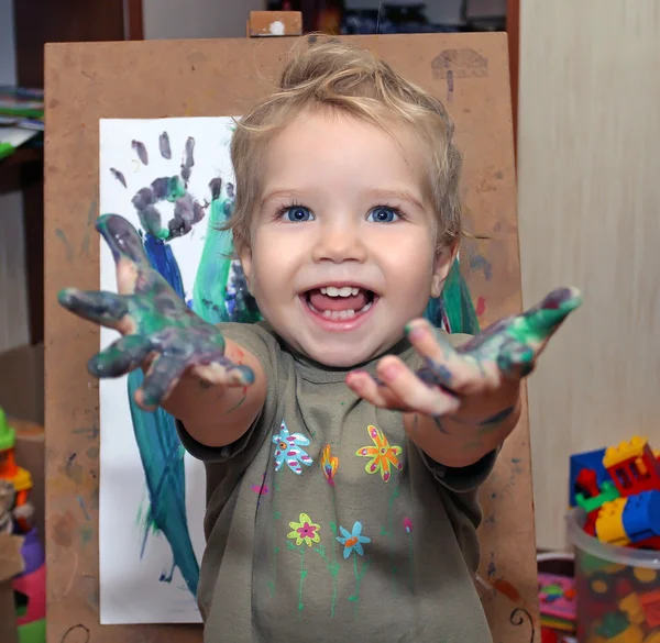 Niño pequeño con ojos azules, dibujando pinturas para los dedos. Babe muestra las palmas manchadas con una pintura. Pequeño pintor con un caballete pintando la hierba . — Foto de Stock