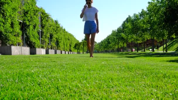 Kvinnan går omkring i det frodiga gräset och pratar i telefon. Kvinna klädd i vit t-shirt och blå shorts går nakna fötter, genom en grön gräsmatta. Frihet och njuta av livet på en solig dag. Stockfilm