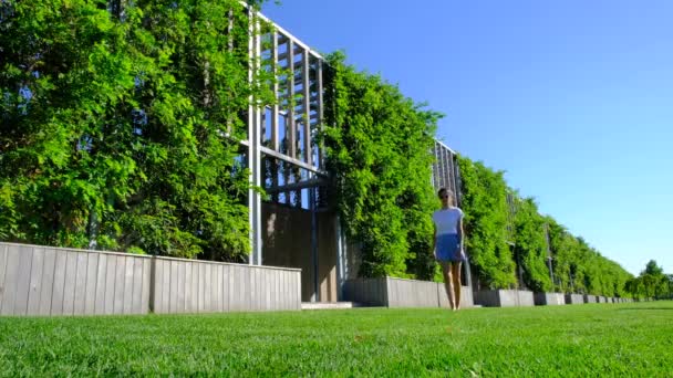 Female wearing in white t-shirt and blue shorts walking bare feet, through a green lawn. Woman walking around garden in park on the lush grass. Freedom and enjoying life on a bright sunny day. — Vídeo de Stock