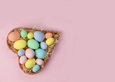 Pastel, pembe arka planda kalp şeklinde bir sepette çok renkli Paskalya yumurtaları. Üst görünüm, metin için yer. Tebrik kartı.