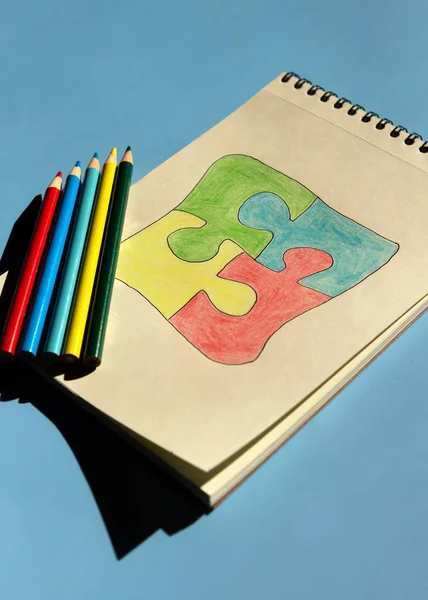 Autism awareness day or month. The drawn puzzle is colored in different colors as a symbol of autism awareness on a blue background. Sketchbook and pencils for children's creativity.