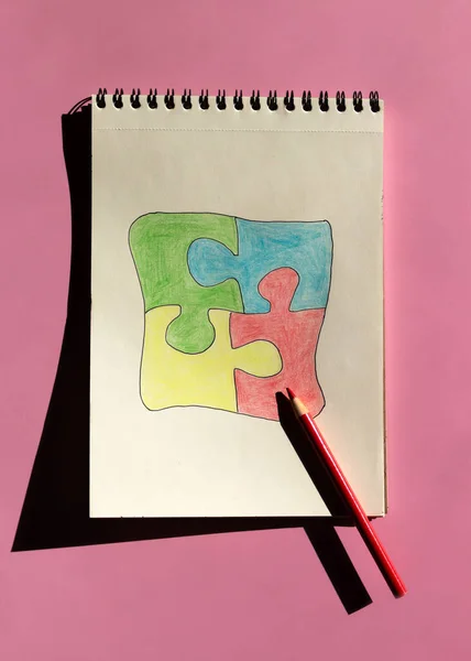 Autism awareness day or month. The drawn puzzle is colored in different colors as a symbol of autism awareness on a pink background. Sketchbook and pencils for children\'s creativity.