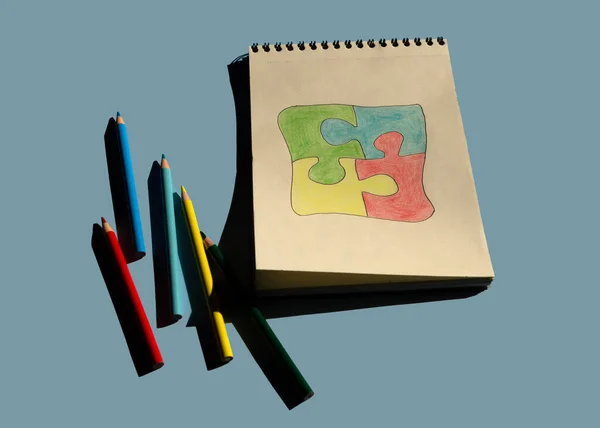 Autism awareness day or month. The drawn puzzle is colored in different colors as a symbol of autism awareness on a blue background. Sketchbook and pencils for children\'s creativity.