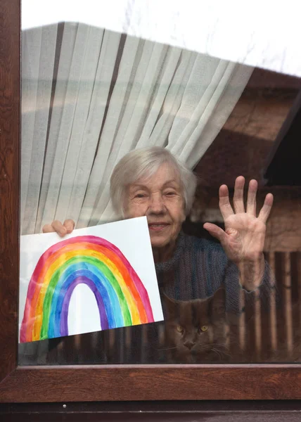 Senior woman in the window of the house holds a childs drawing of a rainbow in her hands. Rainbow in the window.Thank you NHS, Promotion in support of doctors in the coronavirus pandemic, covid 19.