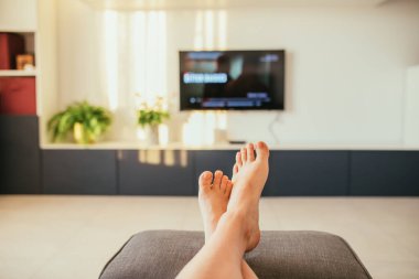 Modern bir oturma odasında doğal gün batımı ışıklarıyla rahatlayan ayakların ve televizyonun seçici odak noktası.. 