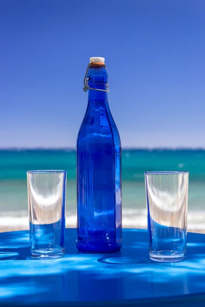 Blue bottle and two glasses on blue table against sea — Stock Photo, Image