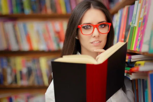 Mujer joven con gafas leyendo cerca de la estantería —  Fotos de Stock