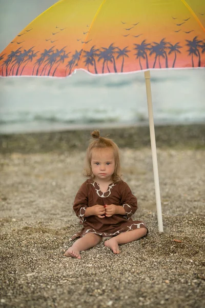 ビーチで傘の下に座っているダウン症候群の美しい女の子 — ストック写真