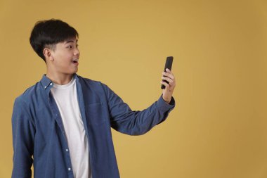 Stüdyonun arka planında izole edilmiş akıllı telefona bakarak sıradan giyinen mutlu Asyalı genç adam portresi.