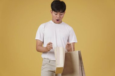 Stüdyonun arka planında izole edilmiş günlük alışveriş torbalarını açan heyecanlı Asyalı genç adamın portresi.