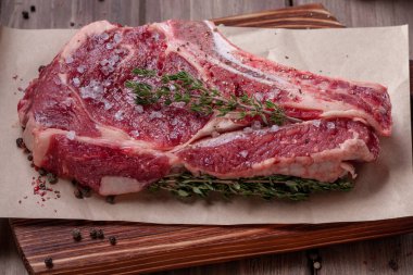 Çiğ pişmemiş biftek. Kemiğin üzerinde büyük bir parça et, baharatlar ve taze otlarla birlikte ahşap bir tahtanın üzerinde yatıyor. Pişirmeye hazır. Etobur ya da ketojenik diyet.