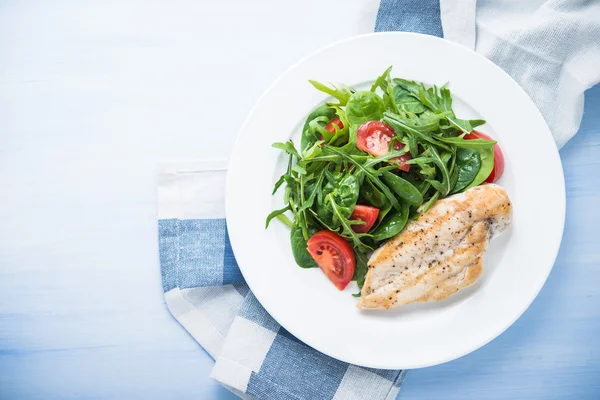 Roasted chicken breast and fresh salad with tomato and greens (spinach, arugula) top view on blue wooden background