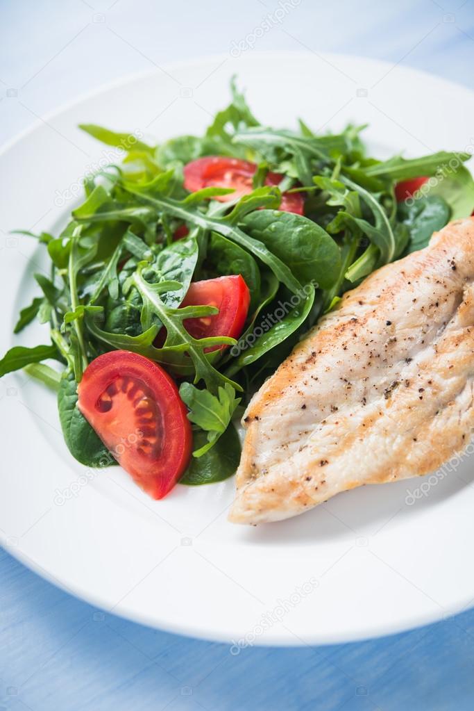 Roasted chicken breast and fresh salad with tomato and greens (spinach, arugula) close up on blue wooden background