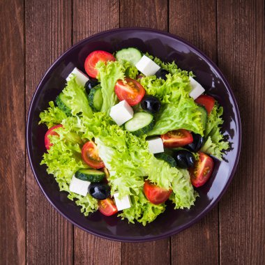 Yunan salatası (marul, domates, beyaz peynir, salatalık, siyah zeytin) koyu ahşap arka plan üst görünümünde.
