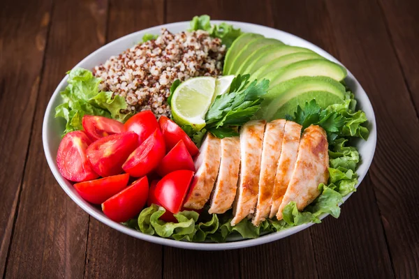 Healthy salad bowl with quinoa, tomatoes, chicken, avocado, lime and mixed greens (lettuce, parsley) on wooden background close up.