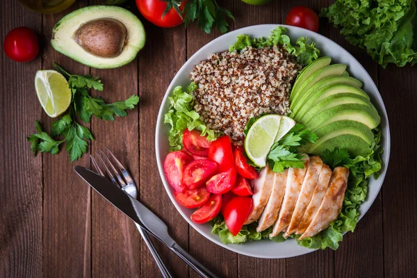 Ahşap arka plan üst görünümünde Quinoa, domates, tavuk, avokado, limon ve karışık yeşillik (marul, maydanoz) ile Sağlıklı salata kase.