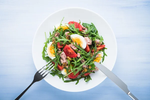 Frischer Salat mit Thunfisch, Tomaten, Eiern, Rucola und Senf auf blauem Holzhintergrund. — Stockfoto