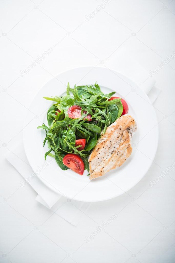 Chicken breast and fresh salad with tomato and greens (spinach, arugula) top view on white wooden background.