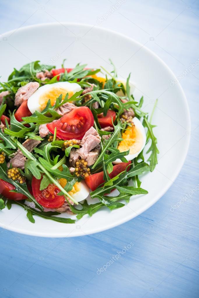 Fresh salad with tuna, tomatoes, eggs, arugula and mustard on blue wooden background close up.